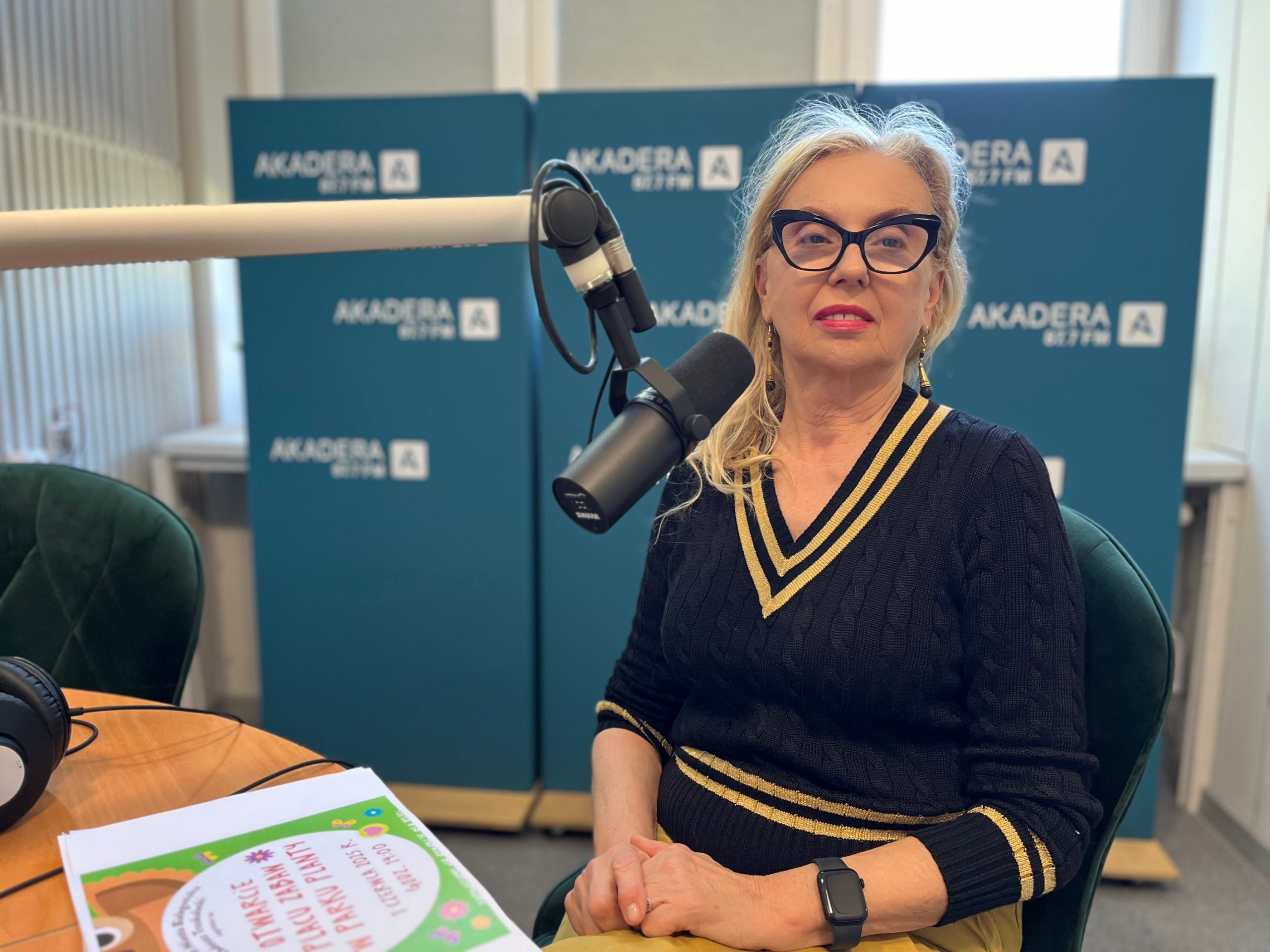 Jolanta Szczygieł-Rogowska, dyrektor Galerii im. Sleńdzińskich, siedzi przy mikrofonie w studiu radiowym. Ubrana jest w czarny sweter z żółtymi akcentami i charakterystyczne okulary. W tle widoczna ścianka z logo Radia Akadera.