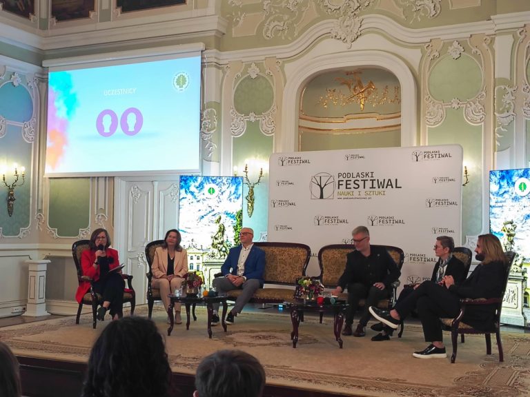 Zabytkowa sala z pięknymi malowidłami, na środku sceny siedzą elegancko ubrani ludzie, trwa debata, w tle logo Dni Nauki i Sztuki i Uniwersytety Medycznego w Białymstoku