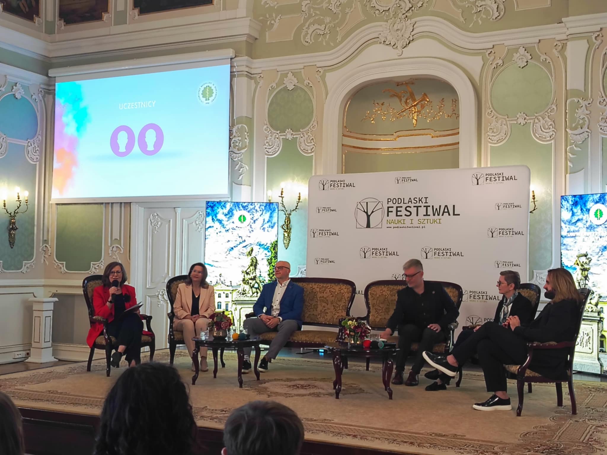 Zabytkowa sala z pięknymi malowidłami, na środku sceny siedzą elegancko ubrani ludzie, trwa debata, w tle logo Dni Nauki i Sztuki i Uniwersytety Medycznego w Białymstoku