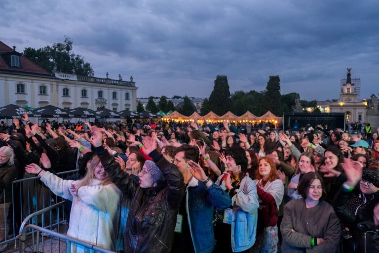 Tłum uśmiechniętych uczestników koncertu na dziedzińcu Pałacu Branickich w Białymstoku. Wieczorna sceneria, sceniczne światła i uniesione ręce świadczą o radosnej atmosferze wydarzenia.