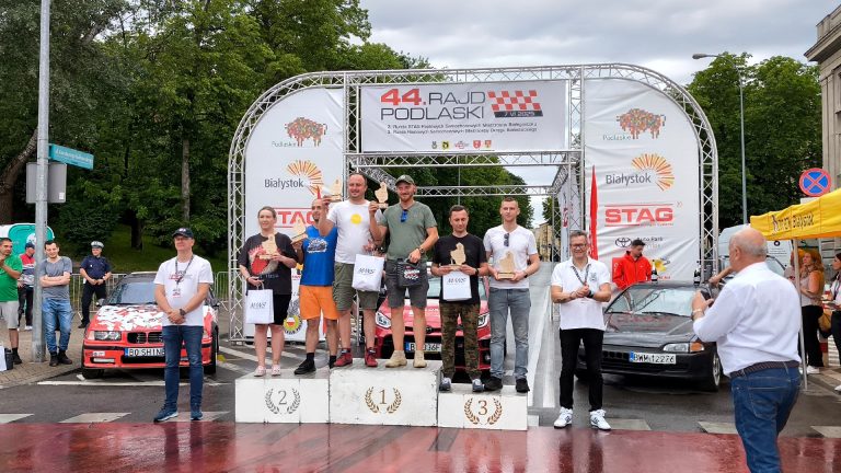 Zwycięzcy 44. Rajdu Podlaskiego stoją na podium podczas ceremonii wręczenia nagród na placu NZS w Białymstoku. W tle baner z logo rajdu oraz sponsorami wydarzenia.