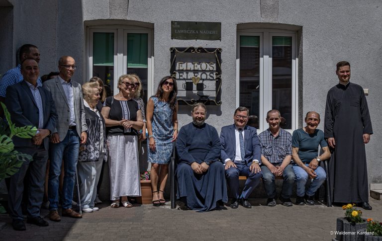 Grupa osób, w tym duchowni, samorządowcy i pracownicy Eleos, pozuje do wspólnego zdjęcia na tle budynku Prawosławnego Ośrodka Miłosierdzia Eleos w Białymstoku. Pośrodku widoczny szyld z napisem „Ławeczka Nadziei” oraz stylizowane metalowe logo Eleos.