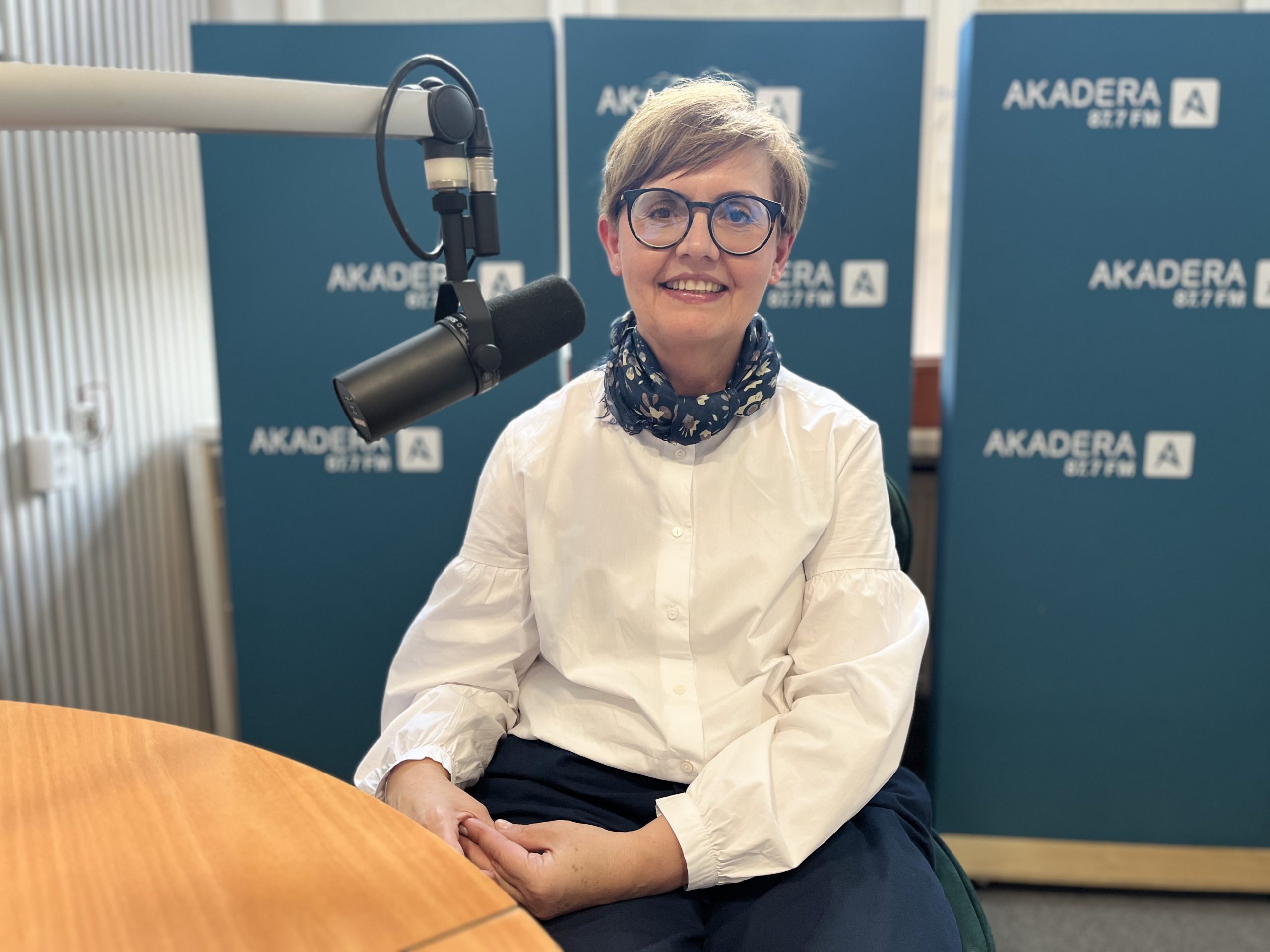 Monika Kordiukiewicz siedzi w studiu Radia Akadera przy mikrofonie, na tle niebieskich plansz z logo stacji. Ma krótkie jasne włosy, okulary i biały koszulowy top.