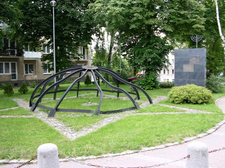 Pomnik upamiętniający Wielką Synagogę w Białymstoku – metalowa konstrukcja w kształcie kopuły oraz czarna płyta z menorą, ustawione na trawiastym skwerze wśród drzew i otaczających bloków.
