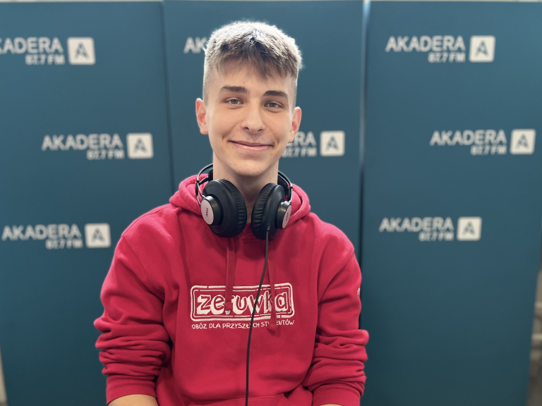 Piotr Tumiel, młody mężczyzna w czerwonej bluzie z logo „Zerówka”, siedzi uśmiechnięty w studiu Radia Akadera, na szyi ma słuchawki. W tle niebieskie ścianki z logo „AKADERA 87.7 FM”.