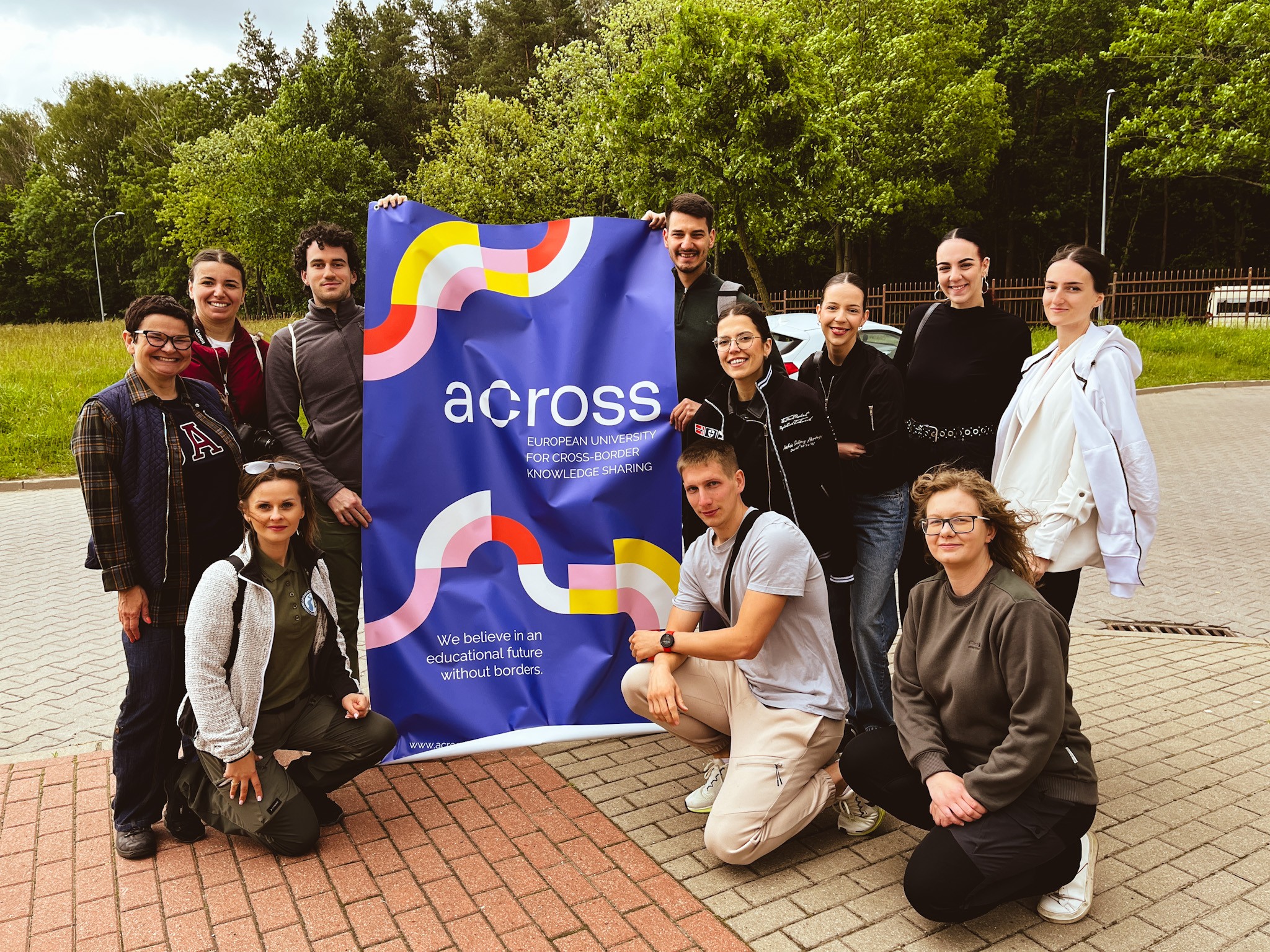 Uśmiechnięta grupa studentów i opiekunów pozuje na tle banera projektu ACROSS. Zdjęcie wykonane na tle zieleni i drzew, przed Silvarium w Poczopku.