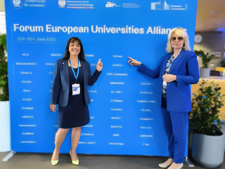 Dwie kobiety w niebieskich marynarkach stoją na tle niebieskiej ścianki z logo III Forum Uniwersytetów Europejskich we Wrocławiu i wskazują na nazwę swojego sojuszu ACROSS.