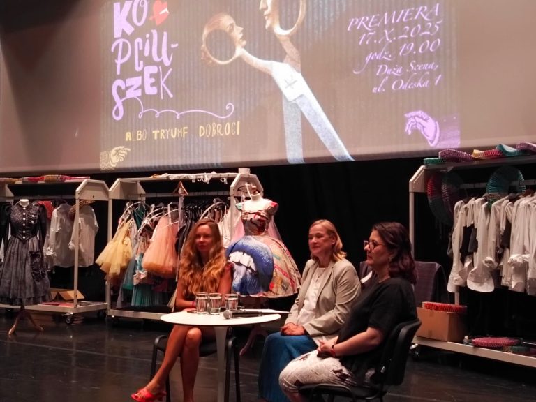 Na zdjęciu widzimy trzy kobiety siedzące na scenie podczas spotkania zapowiadającego spektakl „Kopciuszek albo triumf dobroci”. Za nimi znajdują się wieszaki z teatralnymi kostiumami – suknie balowe, koszule i inne elementy garderoby scenicznej. W tle wyświetlona jest grafika promująca premierę przedstawienia, która odbędzie się 17 października 2025 roku o godz. 19:00 na Dużej Scenie. Scenografia i stroje tworzą barwną, bajkową atmosferę. Zdjęcie oddaje klimat przygotowań i kreatywnej pracy nad spektaklem.