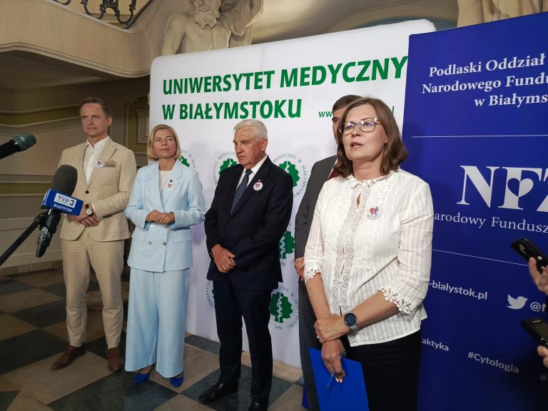 Na zdjęciu widzimy konferencję prasową zorganizowaną wspólnie przez Uniwersytet Medyczny w Białymstoku oraz Podlaski Oddział Narodowego Funduszu Zdrowia. Przed ściankami z logotypami instytucji stoją przedstawiciele organizatorów, m.in. kobieta w białej bluzce, mężczyzna w jasnym garniturze oraz kobieta w błękitnym garniturze. Wszyscy mają przypięte identyfikatory z grafiką kampanii zdrowotnej, co sugeruje, że wydarzenie dotyczyło profilaktyki, edukacji zdrowotnej lub ogólnopolskiego programu badań. Przed uczestnikami ustawiony jest mikrofon TVP3 Białystok, co wskazuje na obecność mediów.