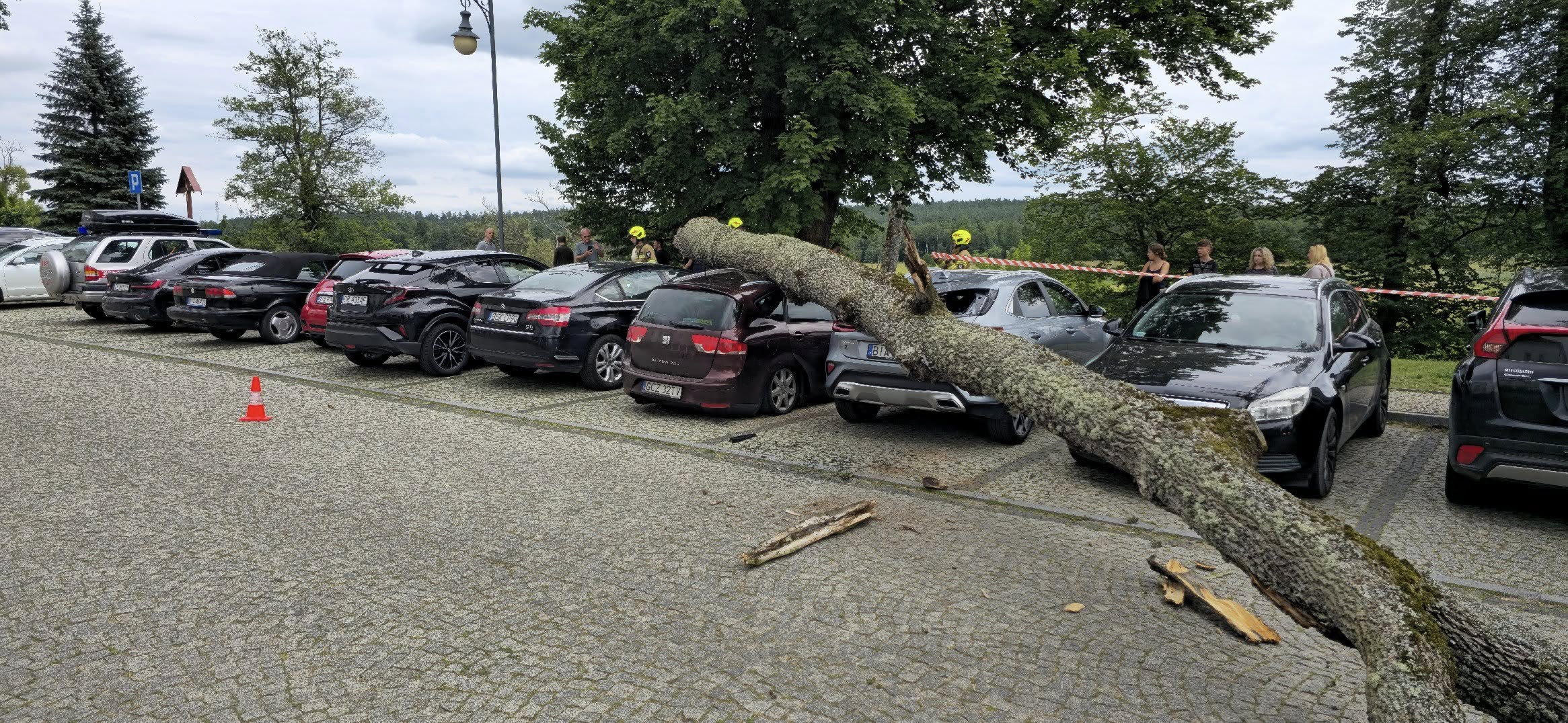 Zawalony pień drzewa leży na zaparkowanych samochodach w Supraślu, w tle ludzie i taśma zabezpieczająca teren.