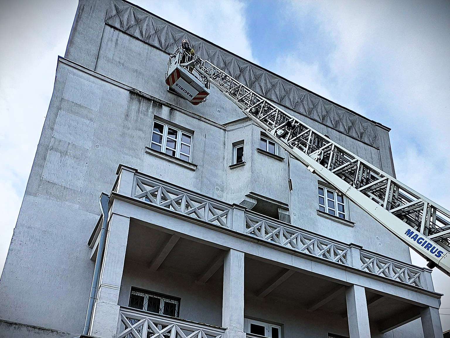 Strażak na wysięgniku drabiny ewakuacyjnej podczas akcji ratowniczej w hostelu przy ulicy Abramowicza w Białymstoku – budynek o jasnej elewacji, niebo z lekkim zachmurzeniem.