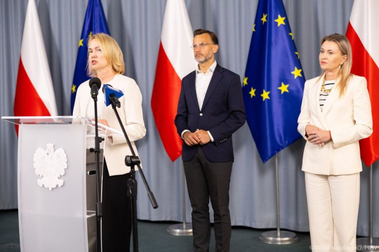 konferencja prasowa stoi mezczyzna i dwie kobiety na tle flag Polski i UE