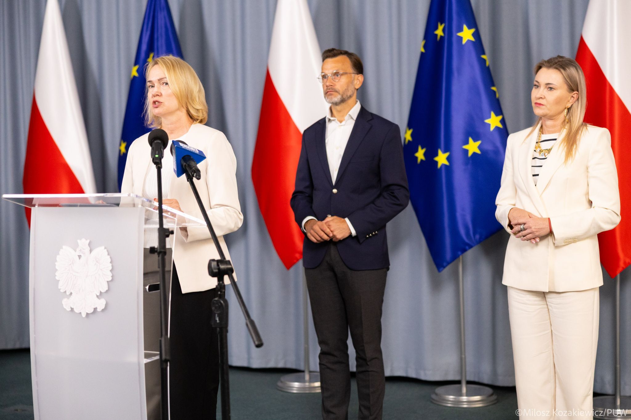 konferencja prasowa stoi mezczyzna i dwie kobiety na tle flag Polski i UE