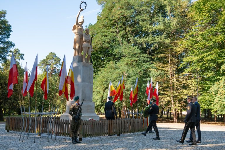 Pomnik żołnierzy 42. Pułku Piechoty otoczony biało-czerwonymi i żółto-czerwonymi flagami w Parku Konstytucji 3 Maja w Białymstoku. Przed pomnikiem stoją żołnierze warty honorowej, a delegacja mundurowych i urzędników składa wieniec z okazji 105. rocznicy Bitwy Białostockiej.