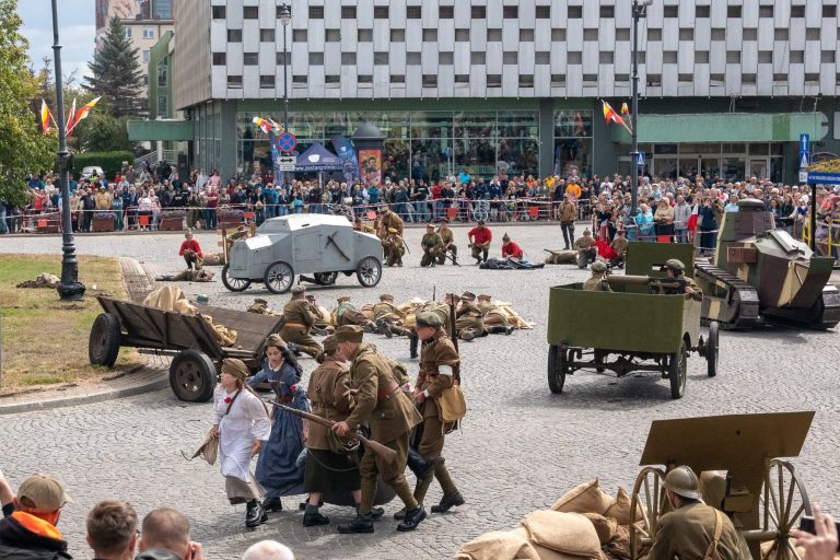 Rekonstruktorzy w historycznych mundurach odtwarzają walki z 1920 roku podczas Bitwy Białostockiej. Na placu NZS w Białymstoku widoczny sprzęt wojskowy i tłumy mieszkańców oglądających widowisko.