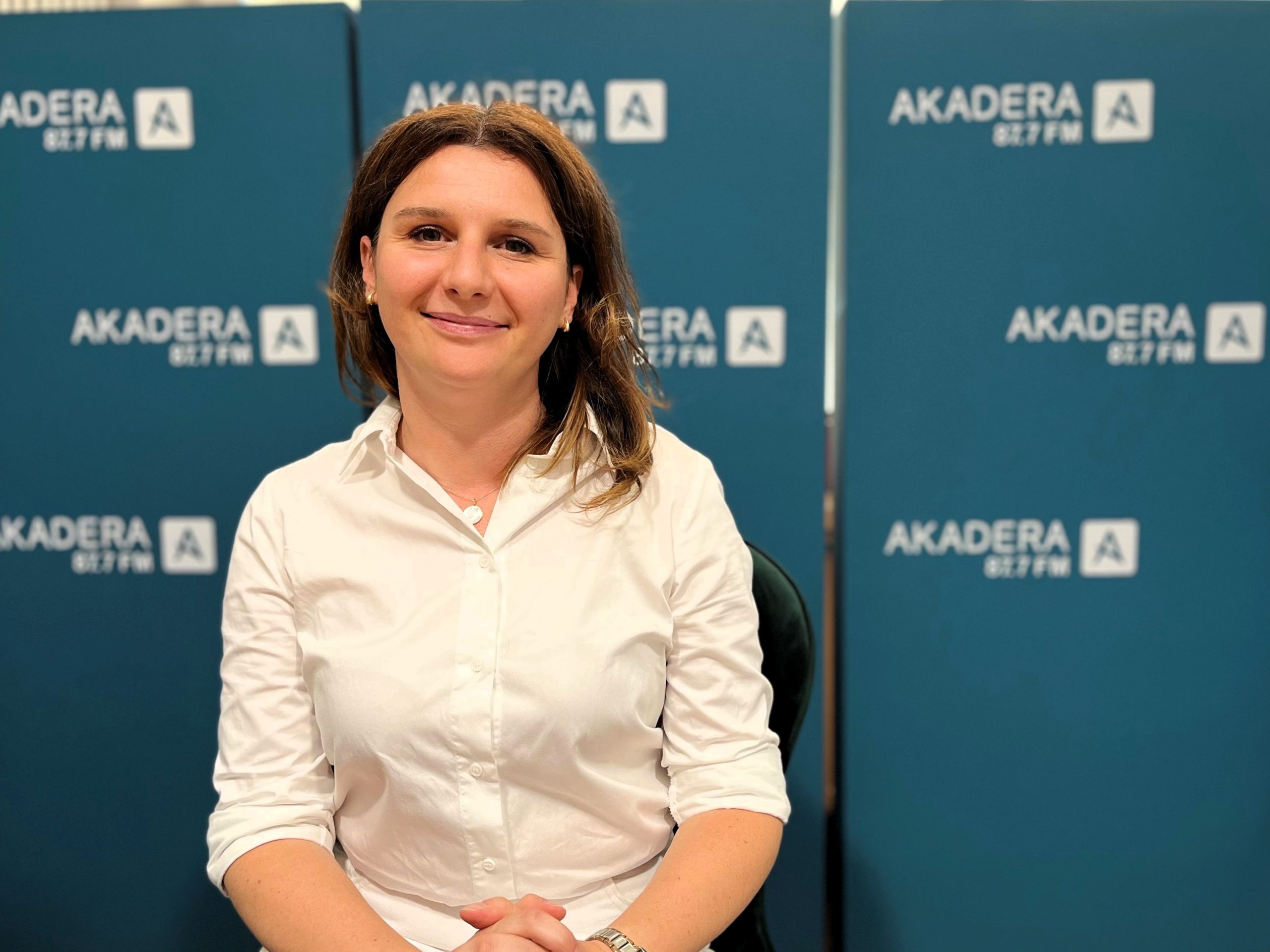 Uśmiechnięta kobieta w średnim wieku, w jasnej koszuli siedzi na tle ścianki z napisem Radio AKADERA