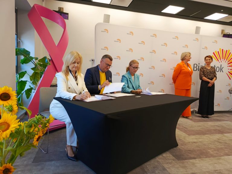 Na zdjęciu widzimy moment podpisania dokumentów w oficjalnym wydarzeniu miejskim w Białymstoku. Trzy osoby — dwie kobiety i jeden mężczyzna — siedzą przy stole, podpisując dokumenty. W tle widoczna jest ścianka z logotypem miasta oraz charakterystyczny różowy symbol walki z rakiem piersi, co może sugerować tematykę zdrowotną lub profilaktyczną. Obecne są również dwie inne kobiety, stojące po prawej stronie, a dekorację sali uzupełniają słoneczniki. Atmosfera jest elegancka i oficjalna.