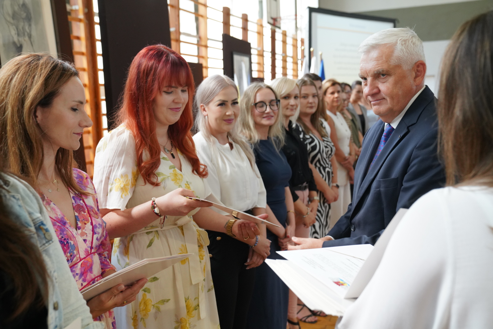 Prezydent Białegostoku wręcza akty mianowania nauczycielom podczas uroczystej ceremonii w IV LO im. Cypriana Kamila Norwida. W tle stoją nauczycielki odbierające dokumenty.