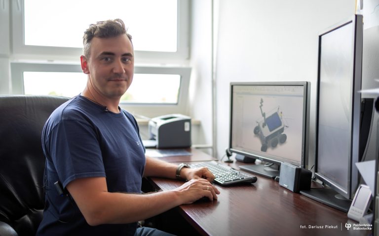 Dr hab. inż. Paweł Dzienis z Politechniki Białostockiej siedzi przy biurku przed komputerem, na monitorze widoczny jest model robota agrotechnicznego R-BOS.