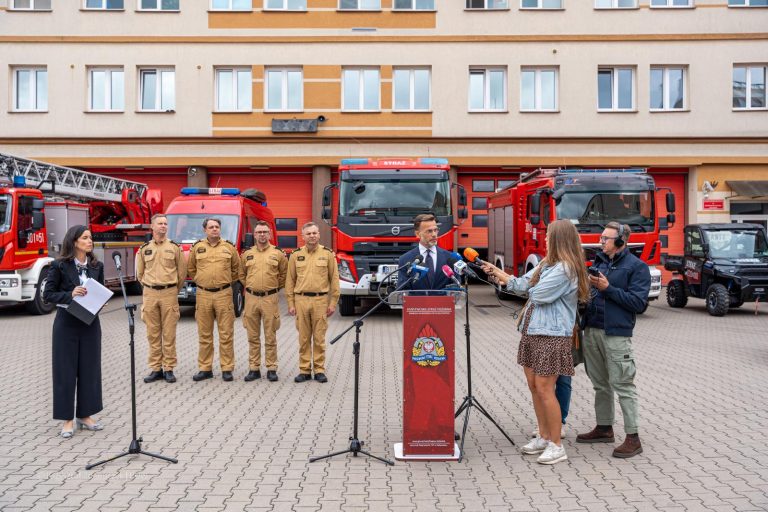 Wojewoda podlaski Jacek Brzozowski przemawia na konferencji przed Komendą Wojewódzką Państwowej Straży Pożarnej w Białymstoku. Obok stoją strażacy w służbowych mundurach, w tle czerwone wozy strażackie i budynek komendy.