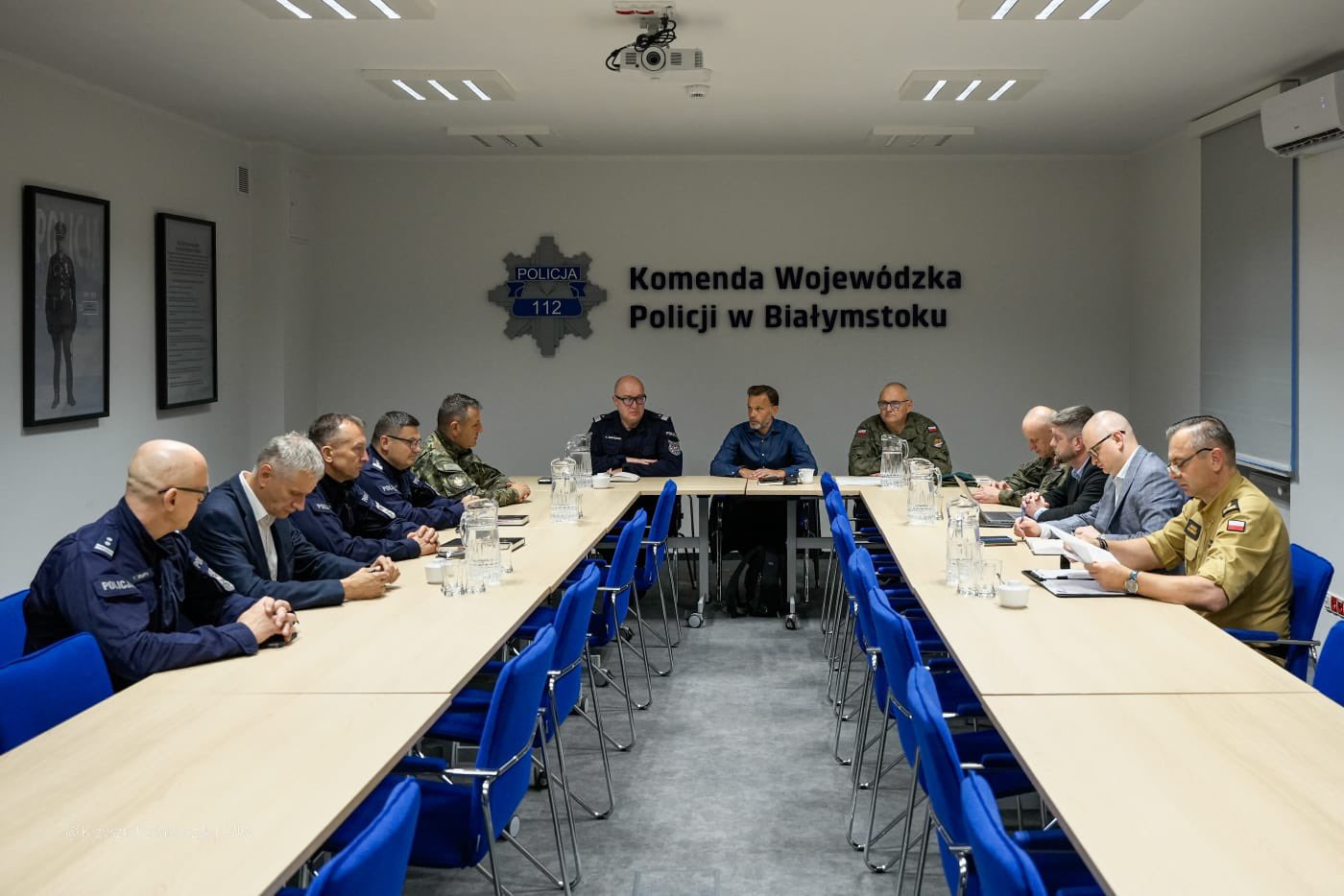 Sala konferencyjna Komendy Wojewódzkiej Policji w Białymstoku. Przy długim stole siedzą wojewoda podlaski, przedstawiciele wojska i policji podczas posiedzenia sztabu kryzysowego.