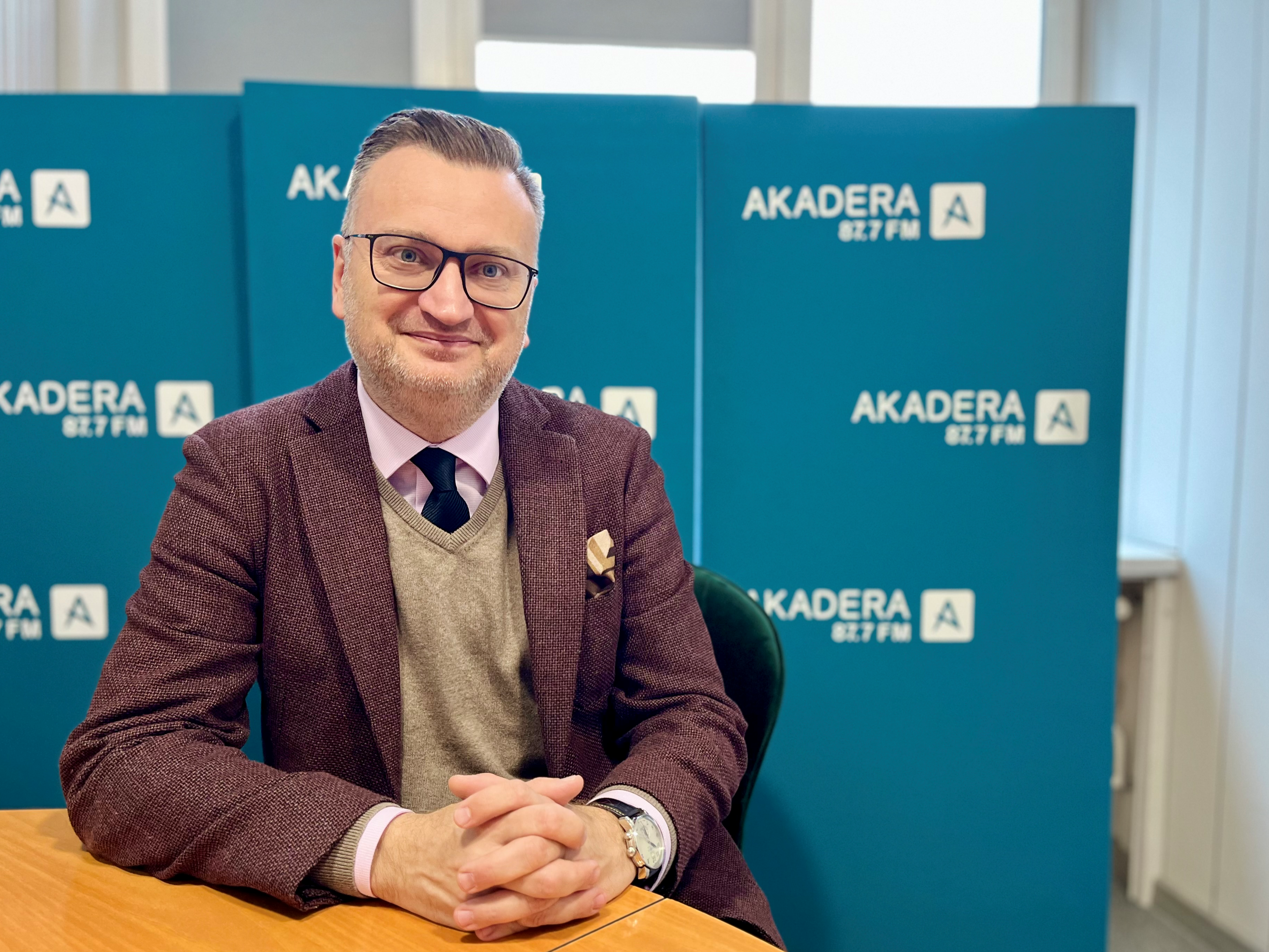 Rafał Rudnicki, zastępca prezydenta Białegostoku, siedzi przy stole w studiu Radia Akadera. Ubrany w bordową marynarkę, szarą kamizelkę i krawat, patrzy w obiektyw i uśmiecha się. W tle niebieskie ścianki z logo Radia Akadera 87.7 FM.