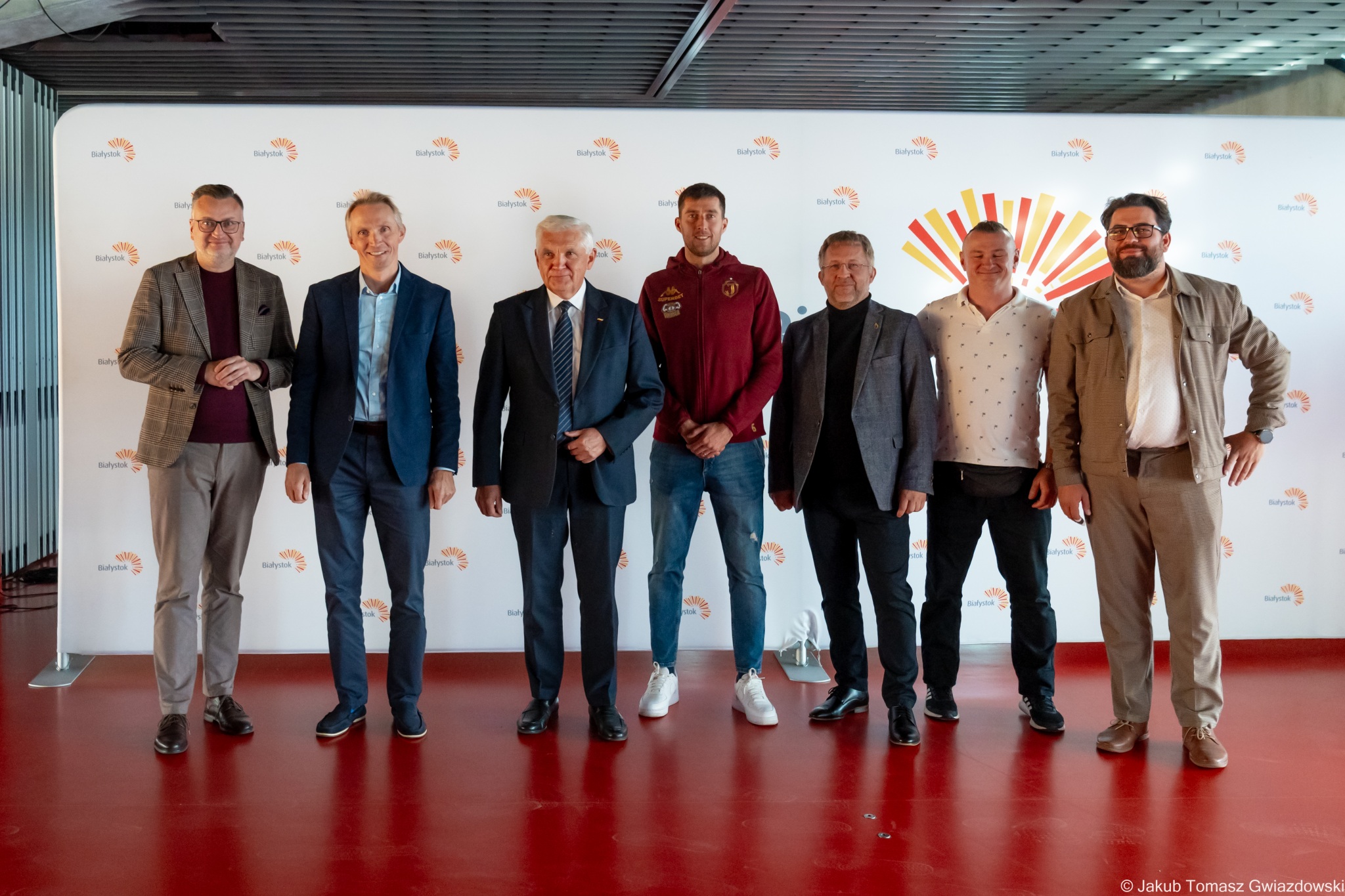 Grupa mężczyzn stoi na tle ścianki promującej Miasto Białystok. Wśród nich przedstawiciele władz miasta, regionu oraz środowiska sportowego podczas prezentacji filmów promujących Białystok i Podlasie w związku z udziałem Jagiellonii w Lidze Konferencji UEFA.