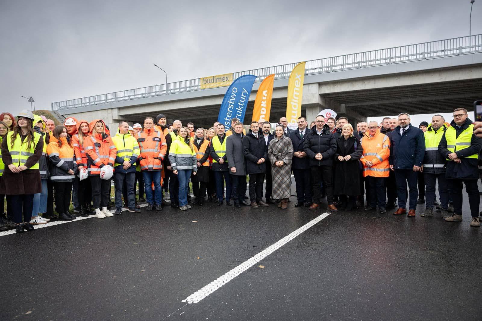 Grupa przedstawicieli Ministerstwa Infrastruktury, wykonawców i samorządowców stoi na nowo otwartym fragmencie drogi ekspresowej S19 w Podlaskiem, w tle widoczny wiadukt i banery firm budowlanych.