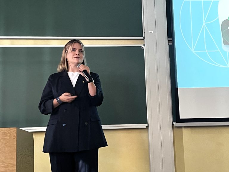 Psycholożka i psychoterapeutka dr Joanna Gutral podczas wykładu o zdrowiu psychicznym na Politechnice Białostockiej, trzyma mikrofon i prezentuje slajd na tle zielonej tablicy.