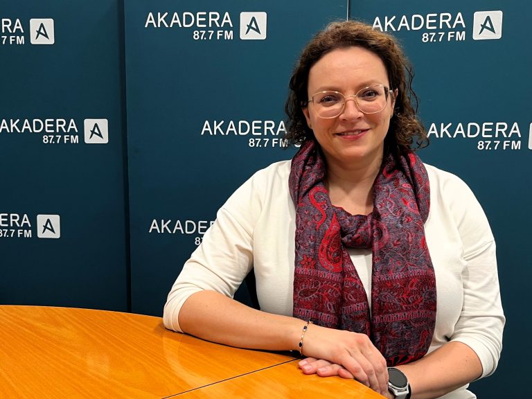 Kobieta z kręconymi włosami i okularami siedzi przy stole w studiu Radia Akadera. W tle widoczne logo „Akadera 87.7 FM”.