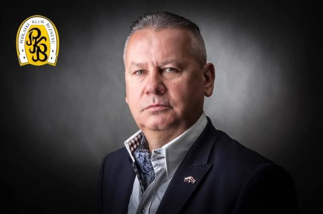 Portret Wojciecha Strzałkowskiego, prezesa Podlaskiego Klubu Biznesu. Mężczyzna w eleganckiej marynarce pozuje na ciemnym tle; w lewym górnym rogu znajduje się logo Podlaskiego Klubu Biznesu.