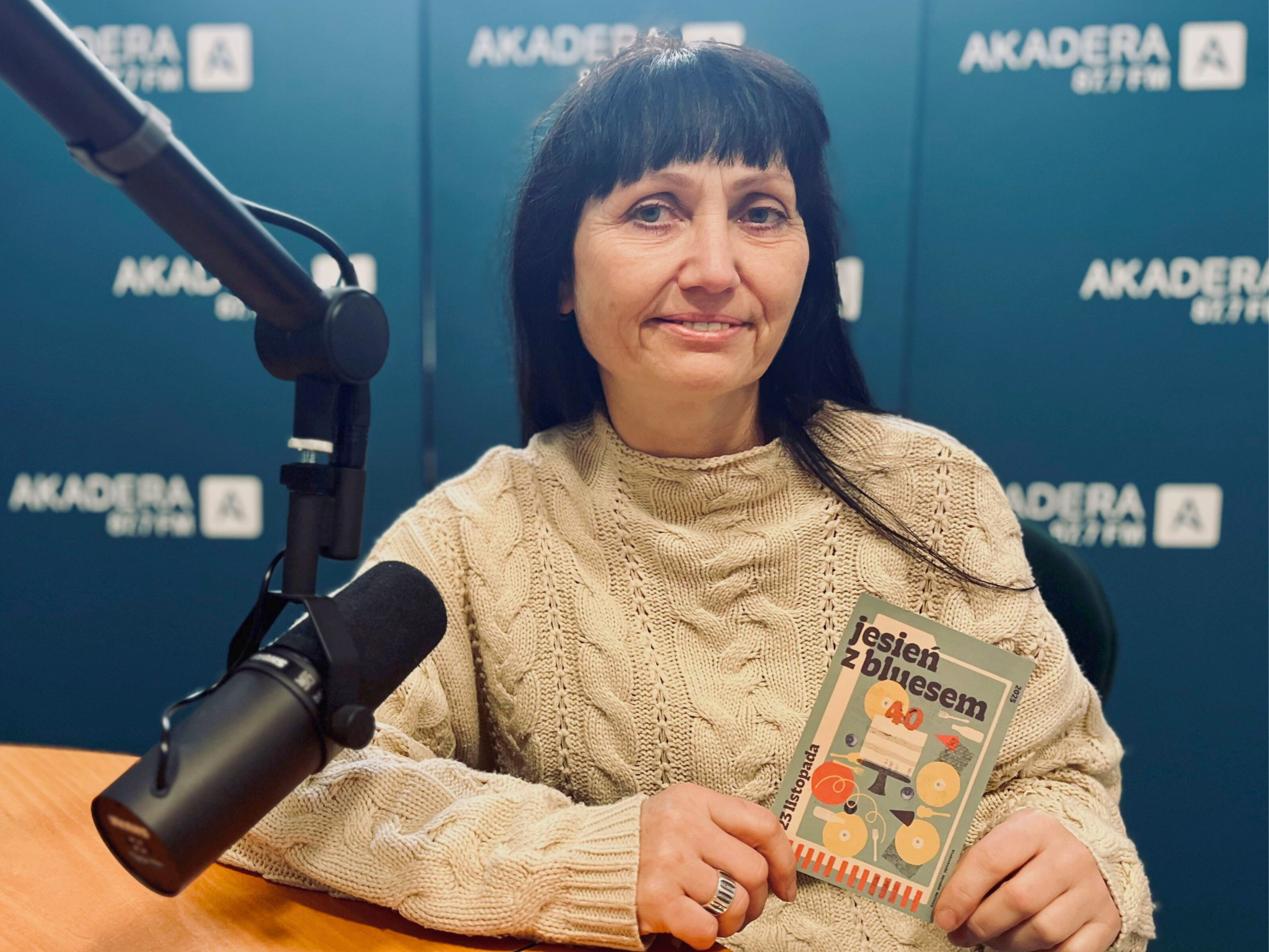 Elżbieta Puczyńska siedzi w studiu Radia AKADERA, trzyma program 40. Jesieni z Bluesem i opowiada o festiwalu. Obok mikrofon i słuchawki na stole, w tle ścianka z logo Radia AKADERA.