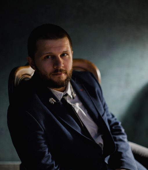Michał Podgórski, prezenter muzyczny Radia AKADERA, siedzi na krześle w eleganckiej marynarce, patrząc w obiektyw w nastrojowym świetle.