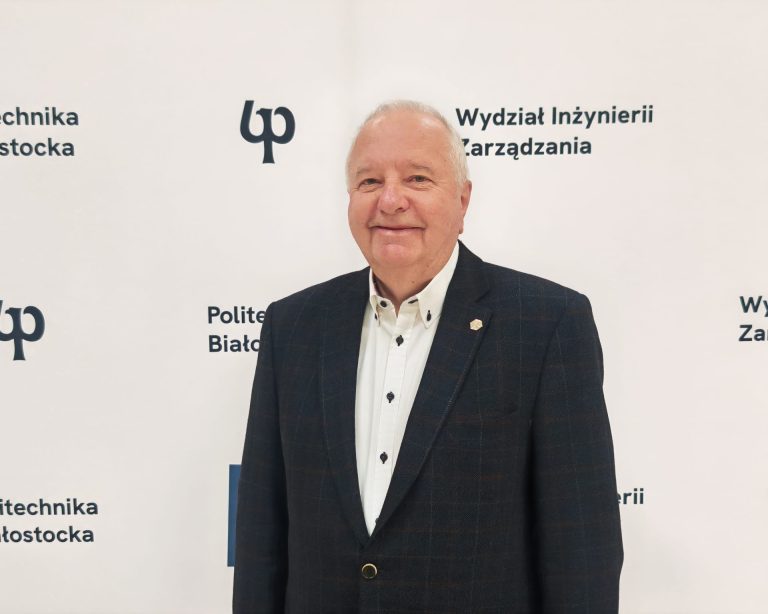 Mężczyzna w marynarce i białej koszuli stoi na tle ścianki Politechniki Białostockiej i Wydziału Inżynierii Zarządzania. To Andrzej Arefiew, przedsiębiorca i właściciel Konsorcjum Arefiew.