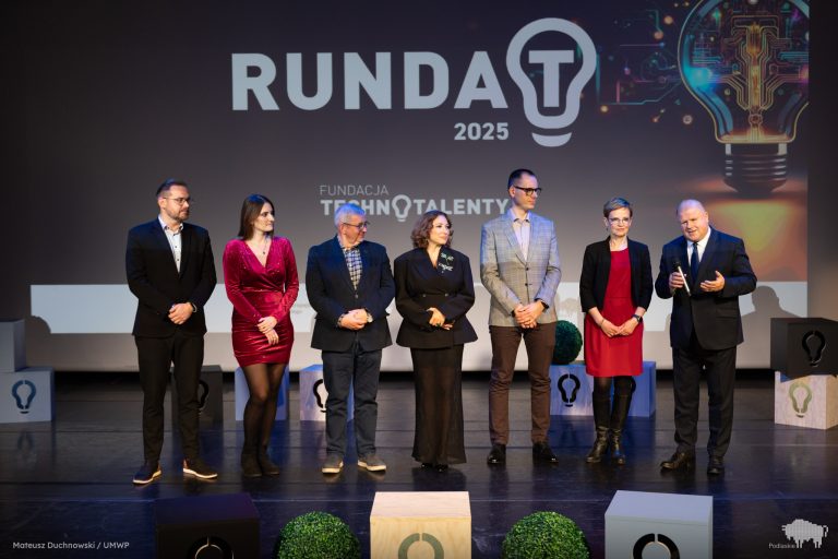Siedmioro uczestników gali konkursu Runda T 2025 stoi na scenie przed dużym ekranem z logo wydarzenia. Wicemarszałek Marek Malinowski przemawia, trzymając mikrofon. Obok niego stoją przedstawiciele Fundacji Technotalenty oraz finaliści konkursu. Tło zdobi grafika żarówki symbolizująca innowacje.